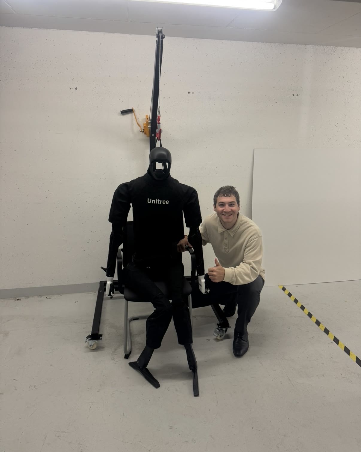 Jordi Spranger crouched next to a black Unitree humanoid robot seated on a test stand, giving a thumbs up.